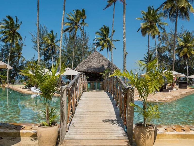 Holzsteg mit Seilen überlauft auf Pool mit Palmen und Strohdachhütte im sonnigen Tropenresort.