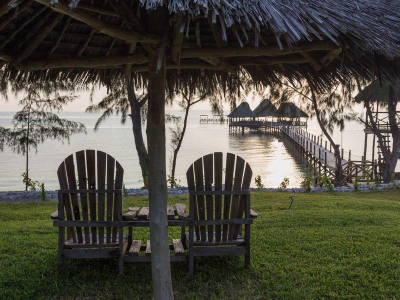 Zwei Holzhocker unter einem Strohdach mit Blick auf Wasser und kleine Hütten am Steg.