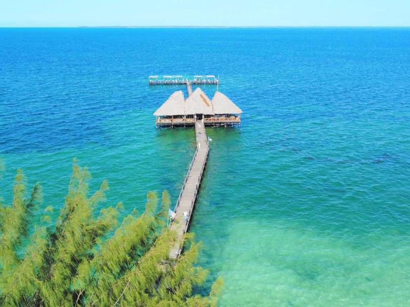 Ein langer Steg führt zu zwei kleinen, über dem Wasser schwebenden Häusern mit Strohdächern.