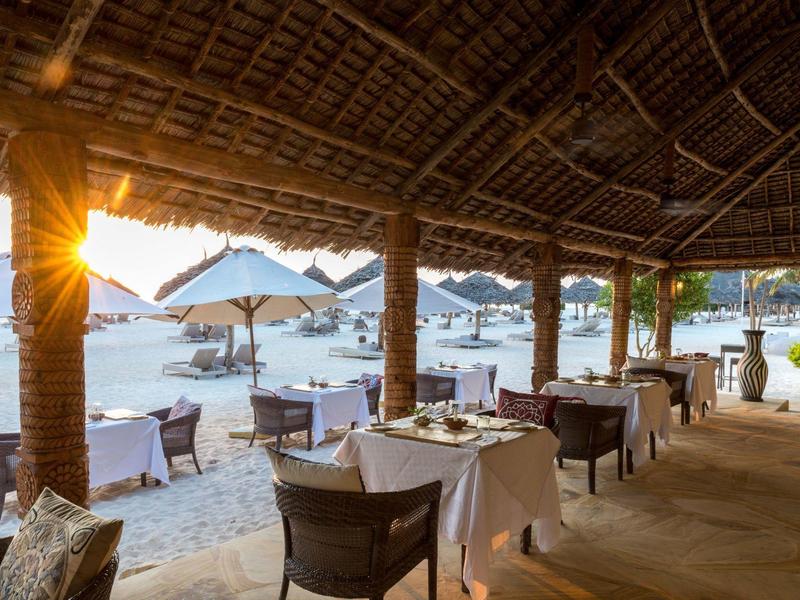 Restaurant mit gedeckten Tischen unter Strohdach am Strand bei Sonnenuntergang.