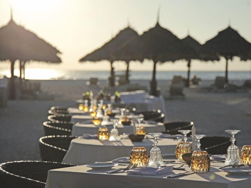 Langer gedeckter Tisch am Strand mit Strohdach-Sonnenschirmen und Sonnenuntergang im Hintergrund.