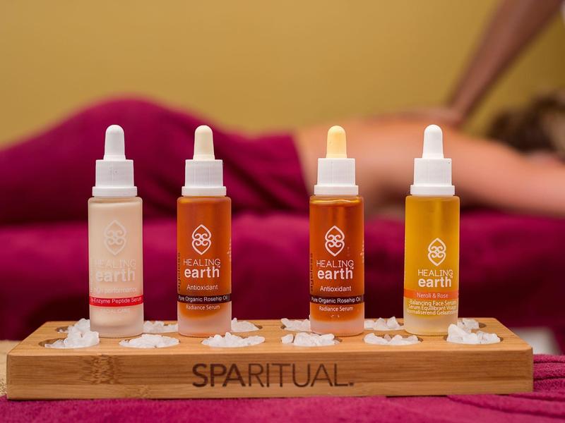 Vier bunte Spa-Ölflaschen stehen auf Holztablett vor liegender, entspannter Person auf rosa Unterlage.