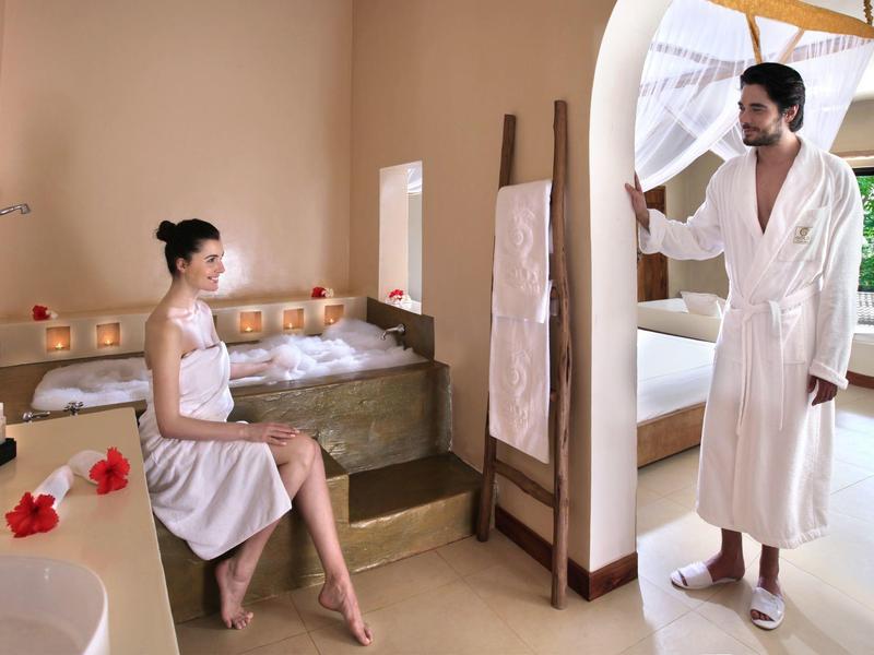 Ein Mann und eine Frau in Bademänteln in einem hellen, modernen Spa-Raum mit Kerzen und Blumen.