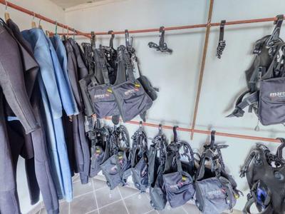 Schwarze und blaue Taucheranzüge hängen an Stangen, dazu mehrere schwarze Taucherwesten an Haken.