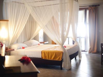 Hell beleuchtetes Schlafzimmer mit Himmelbett, weißen Vorhängen und gelber Decke am Fußende.