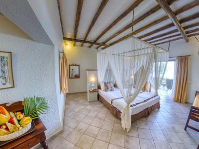 Helles Schlafzimmer mit großen Holzbalken, Himmelbett, Fliesenboden und Obstkorb auf Tisch.