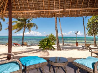 Strand mit Palmen, Sand und blauem Meer, davor Rattanmöbel mit blauen Kissen unter Strohdach.