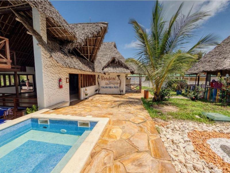 Vrijetijdsgebied van resort met zwembad, rieten bungalows en palmbomen onder blauwe lucht.