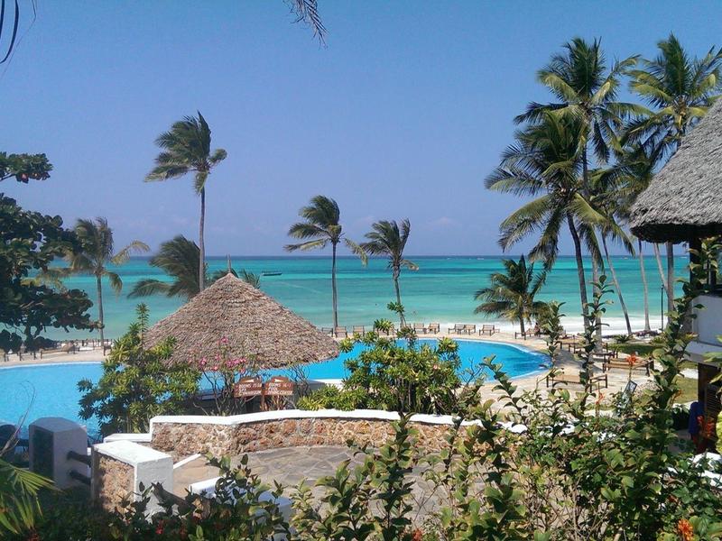 Tropisch resort met zwembad, palmbomen en uitzicht op de blauwe zee onder een heldere lucht.