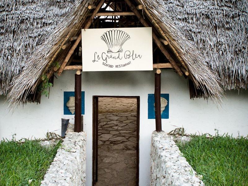 Ingang van een hotel met rieten dak en wit stenen pad omgeven door groen gras.