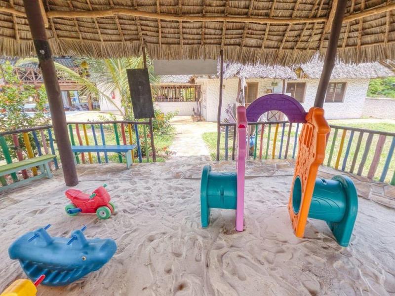 Kinderspeelplaats met glijbaan en zandvloer onder een rieten dak in een vakantiepark.