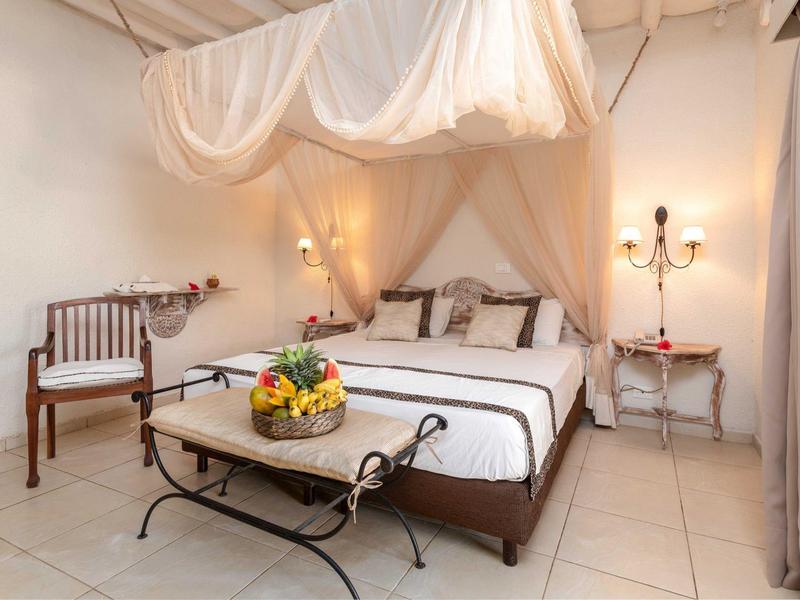 Gezellig tweepersoonsbed met klamboe, houten stoel en fruitmand in lichte hotelkamer.