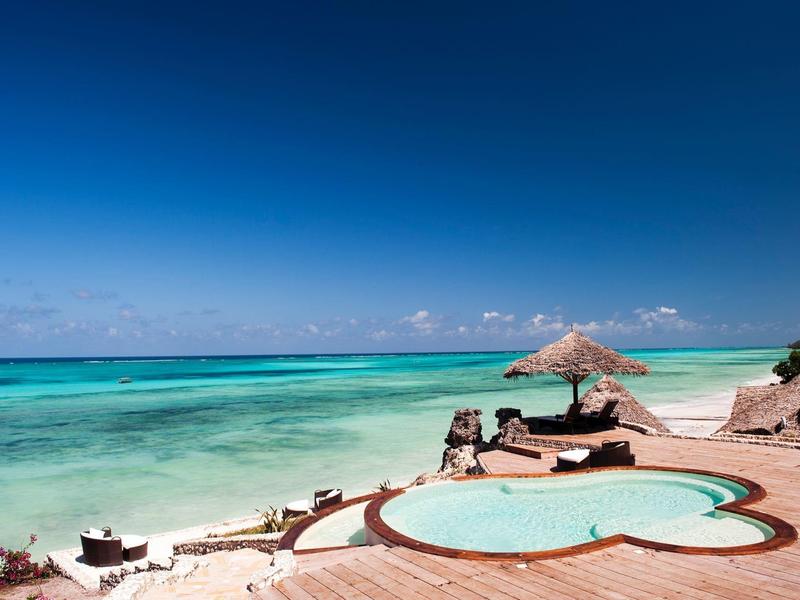 Hotelzwembad met ligstoelen met uitzicht op turquoise zee onder een heldere blauwe lucht.