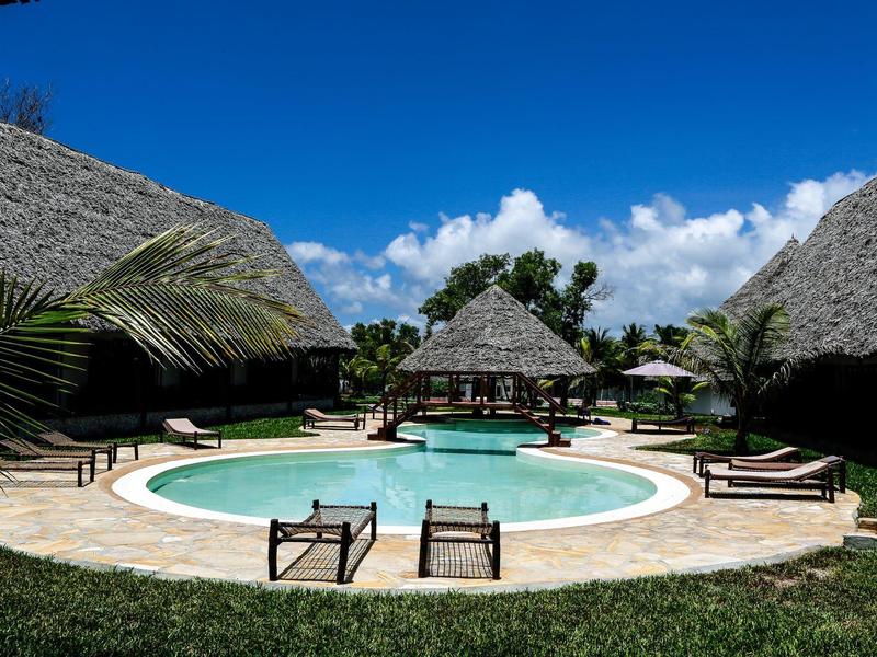 Rond zwembad met ligstoelen en rieten bungalows bij een stralend blauwe lucht.