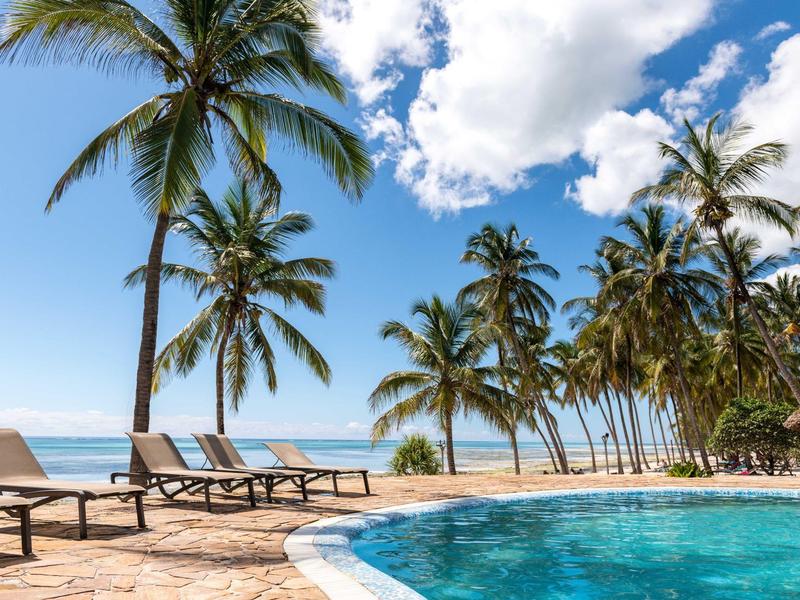 Zwembad met ligbedden en palmbomen aan zee onder een blauwe lucht