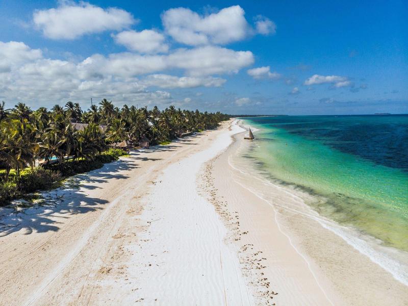 Lange witte zandstrand met palmbomen en helder turquoise water onder een blauwe lucht.