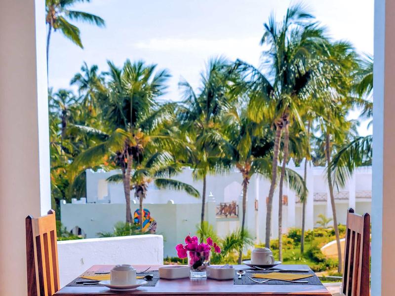 Een gedekte ontbijttafel op een terras met uitzicht op palmbomen en een tuin.
