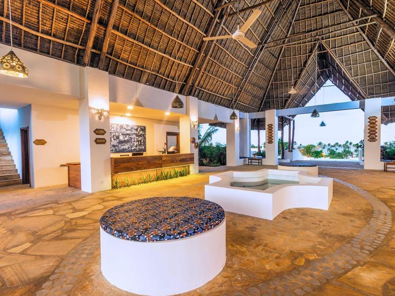 Ruime open hotellobby met rieten dak, zitgedeeltes en receptie bij daglicht.