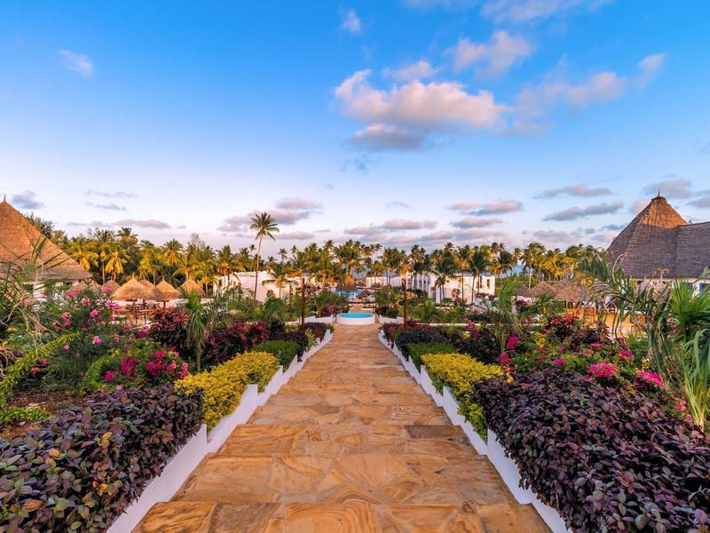 Steenpad door tropische tuin naar een resort met palmbomen en villa's.