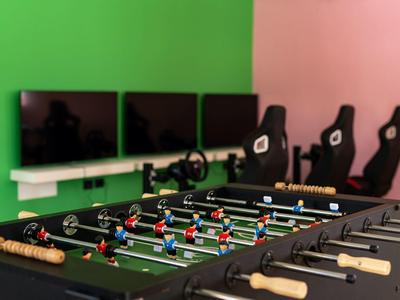 Tischkicker im Vordergrund, dahinter vier Monitore und drei Gaming-Stühle vor grün-roter Wand.