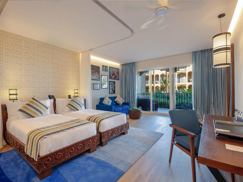 Lichte hotelkamer met twee bedden, blauw tapijt en uitzicht op terras en tuin.