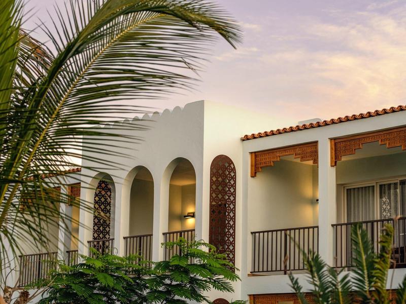Wit hotelgebouw met bogen en balkons omringd door palmbomen bij zonsondergang.