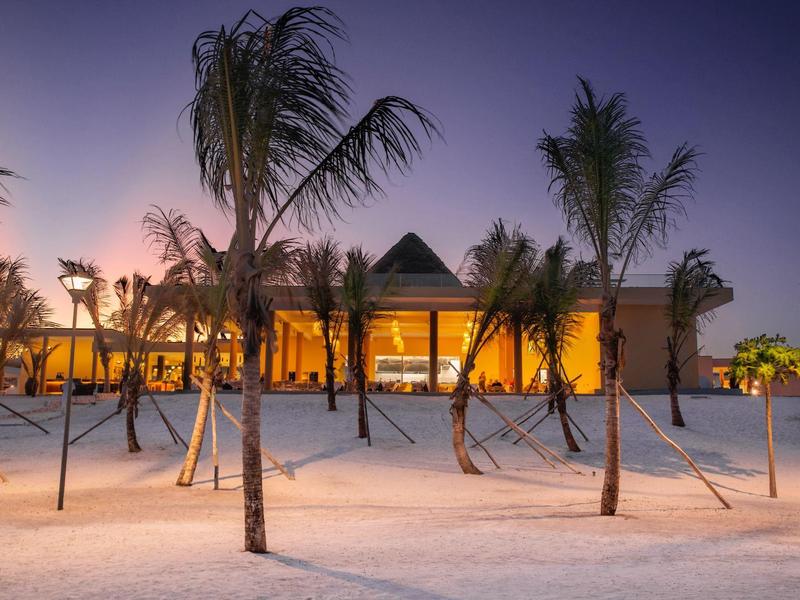 Een tropisch resort bij zonsondergang met slanke palmbomen voor een verlicht gebouw.