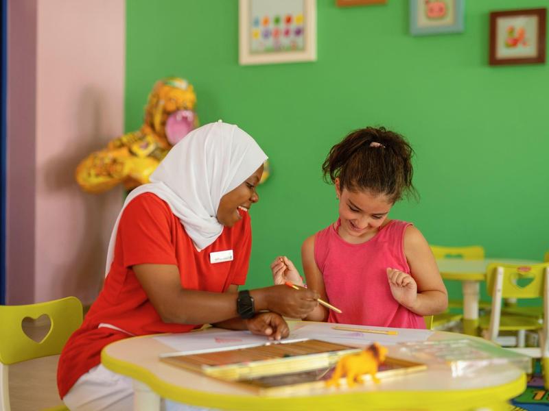 Een begeleider helpt een jong meisje met knutselen in een kleurrijke speelkamer.