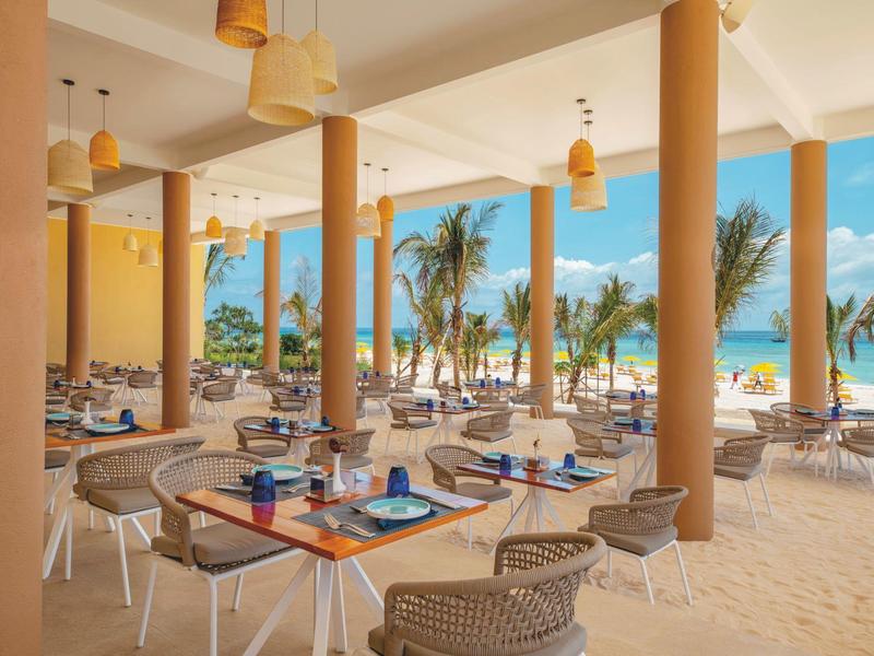 Open strandrestaurant met tafels, stoelen en uitzicht op palmbomen en zee.