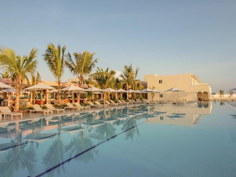 Een rustige hotelzwembad met ligstoelen en palmbomen onder een heldere lucht.