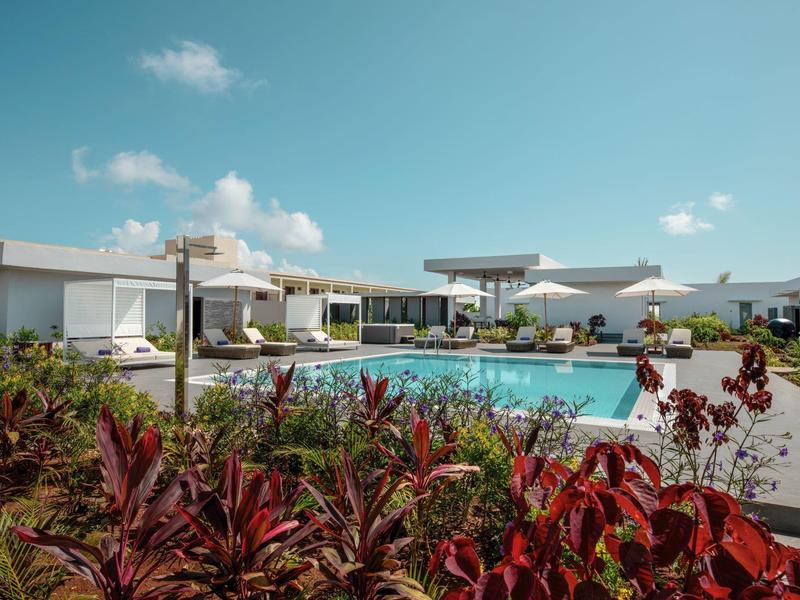 Moderne hotelzwembad met witte schaduwstructuren en tropische planten onder een zonnige lucht.