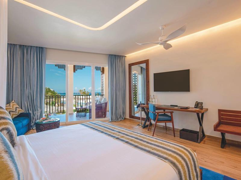 Modern hotelkamer met groot bed, tv, bureau en balkon met uitzicht op zee.