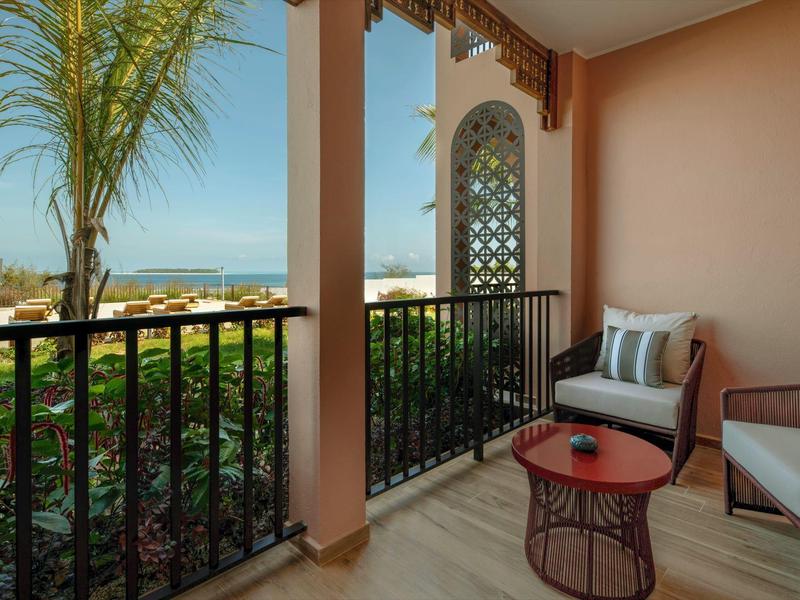Balkon met stoelen en tafel met uitzicht op tuin en zee onder heldere hemel.