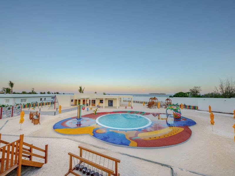 Kleurrijke kinderspeeltuin met plonsbad en zand, omgeven door parasols op het strand.