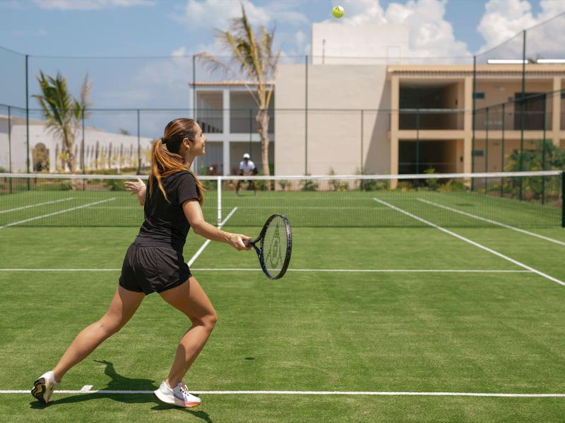 Vrouw speelt tennis op een groene baan in een zonnig buitengebied van een hotel.