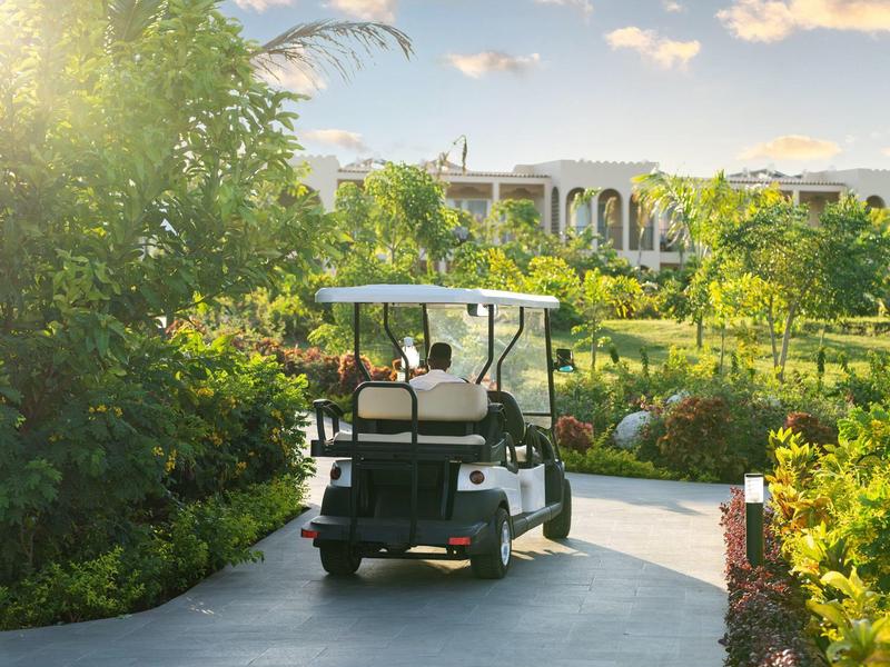 Golfkar rijdt op een pad door een zonnige, groene hotel tuin.