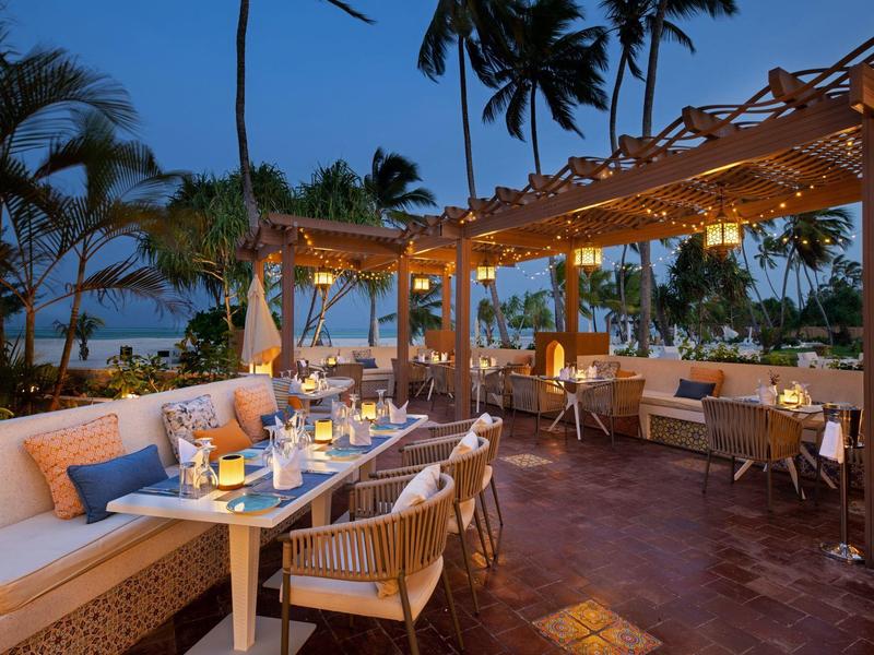 Terraza al atardecer con mesas y sillas bajo una pérgola iluminada en un entorno tropical.