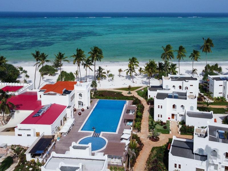 Vista de un hotel con piscina, edificios blancos y palmeras junto al mar de agua azul.