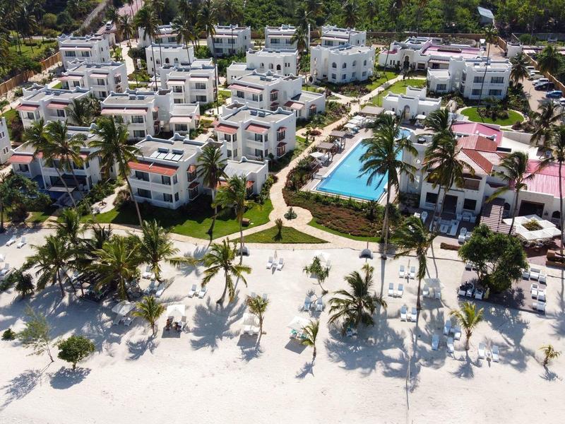 Resort de playa con edificios blancos, piscina y palmeras en la arena bajo cielo azul.