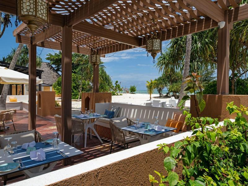 Área de restaurante al aire libre con techo de madera, mesas, sillas y vegetación tropical bajo el sol.