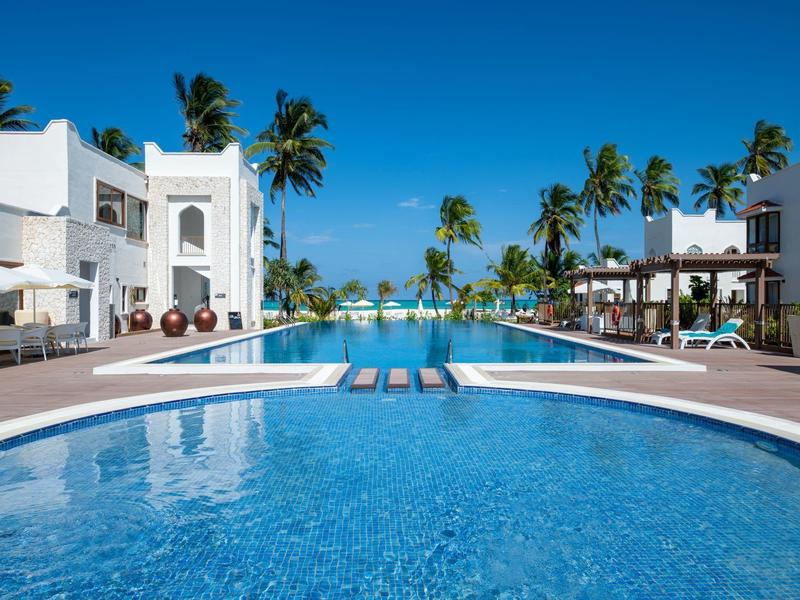 Gran piscina rodeada de edificios blancos y palmeras bajo un cielo azul claro en un resort.
