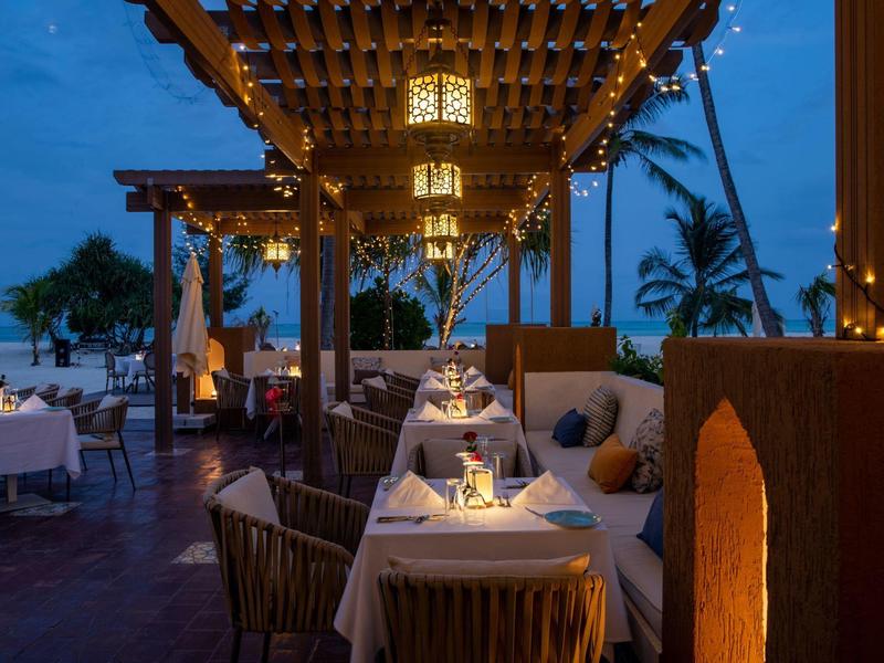 Restaurante al aire libre iluminado con mesas preparadas para la cena en un entorno tropical al atardecer.