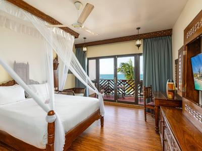 Helles Schlafzimmer mit Himmelbett, Holzboden, Fernseher und Balkon mit Meerblick und blauen Vorhängen.