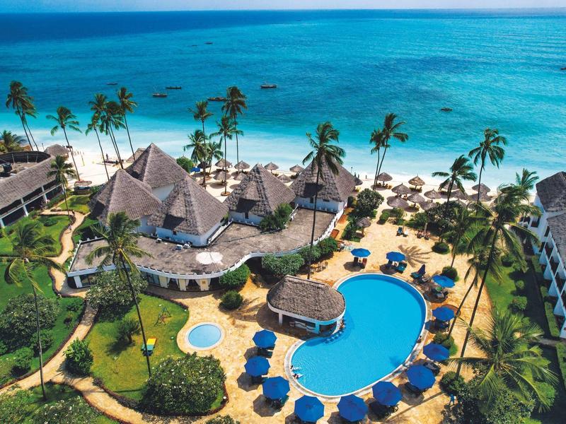 Uitzicht op tropisch hotelresort met zwembad, palmbomen en blauwe zee aan het strand.