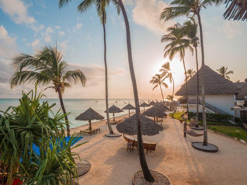 Een tropisch strand met palmbomen, parasols en rustige zee bij zonsondergang.