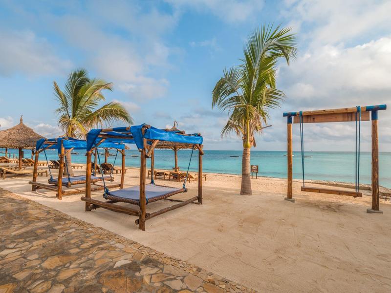 Strand met palmbomen, schaduwrijke ligbedden en een schommel bij zee onder een blauwe hemel.