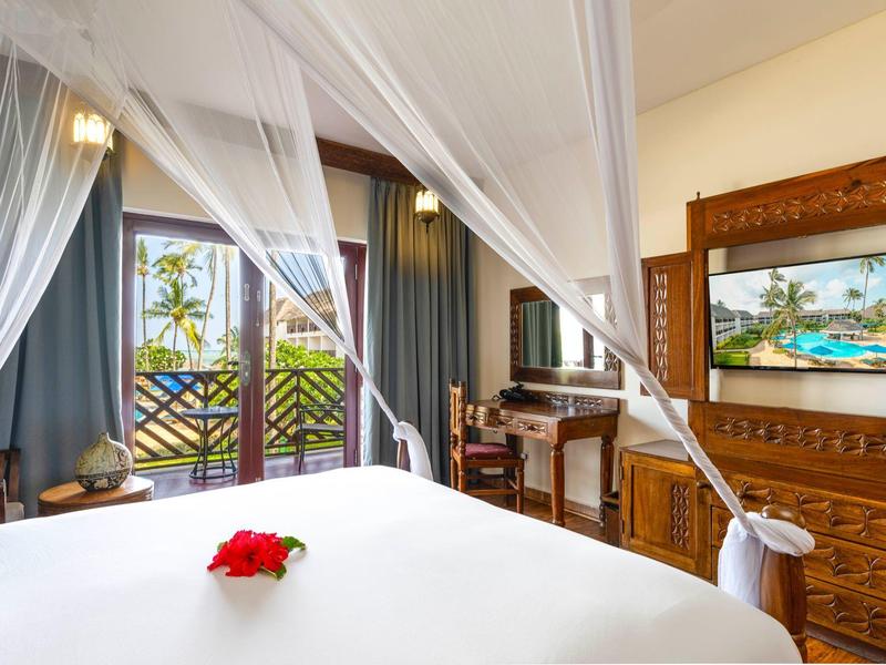 Lichte hotelkamer met hemelbed, balkon en uitzicht op palmbomen en zee.