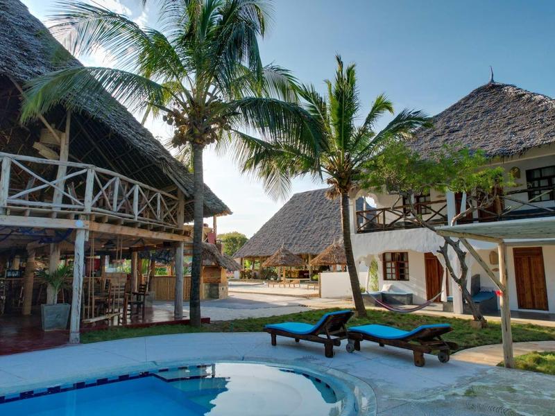 Area piscina con lettini, palme e edifici tradizionali con tetto di paglia sotto un cielo sereno.