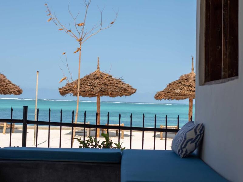 Vista da una terrazza con cuscini su sabbia bianca, ombrelloni di paglia e mare turchese.