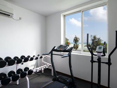 Petite salle de sport avec haltères, tapis roulant et vélo elliptique devant une fenêtre avec vue sur le ciel.